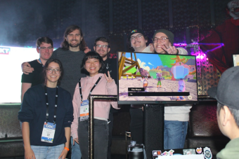 A group of six individuals, pose smiling in front of a display at GDC 2023. They stand behind a monitor showing a colorful game scene with animated characters, including a figure labeled 'The Mayor.' The group includes team members wearing GDC badges and casual attire. The event setting features vibrant lighting and a partially visible red decoration resembling a clown.