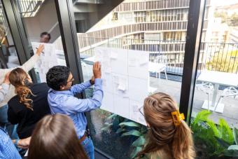 A group of people participating in a design thinking workshop. They are gathered around large sheets of paper with grids and sticky notes, posted on glass windows in a bright, modern building. The background shows an outdoor terrace with white tables and chairs, and a view of contemporary architecture.