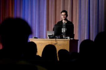 A speaker stands behind a wooden podium, delivering a presentation to an audience in a dimly lit auditorium. The stage backdrop consists of softly lit curtains in shades of blue and red. The audience is visible in the foreground as silhouettes, while the presenter, dressed in black and wearing glasses, speaks into a microphone.