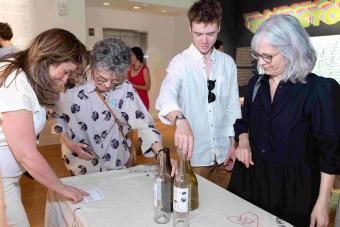 Four visitors gather around an embroidered tablecloth featuring stitched questions and designs, including “BE MINE” in a red heart. One person points to a label, while another places a bottle among others on the table. The colorful “TRAJECTORIES” title is visible on the exhibition wall behind them.