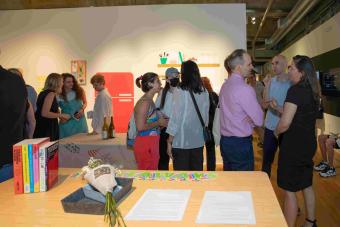 A lively gallery reception scene with attendees mingling and conversing in small groups. In the foreground, a table displays a bouquet wrapped in newspaper, several colorful books, and handouts labeled “TAKEAWAYS.” The background features a brightly lit wall with kitchen-themed illustrations.