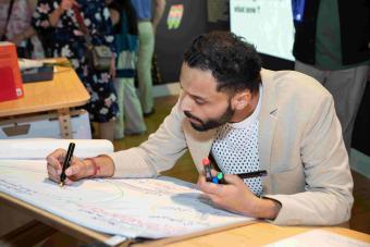 A person in a tan blazer leans over a table and writes on a large sheet of paper with colorful markers, contributing to a collaborative drawing or reflection activity. The background shows gallery attendees in conversation and exhibit displays on the wall.