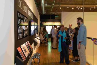 Attendees explore an exhibition titled "Robot Reveals," featuring books, wall text, and interactive displays. A robotic dog rests on the floor near the wall, and people gather in conversation under gallery lighting, with a vibrant “TRAJECTORIES” sign visible in the background.