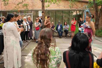 A group of people gathers outdoors in a shaded courtyard for a performance or presentation. One person in the foreground wears a colorful outfit with sculptural accessories and speaks to the audience, while others stand or sit watching attentively under string lights.