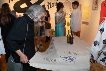 A woman with gray hair wearing a black dress closely examines the embroidered writing on a large textile laid out on a table. Around her, people mingle in a gallery filled with colorful artwork, and three wine bottles rest on the fabric.