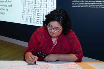 A person in a red floral top writes with colorful markers on a large sheet of paper covered in handwritten notes. Behind them, a black gallery wall displays text and a projected image of a maze.