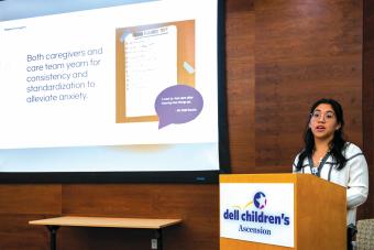 A presenter stands at a podium labeled “Dell Children’s Ascension,” delivering a talk in front of a projection screen. The slide reads, “Both caregivers and care team yearn for consistency and standardization to alleviate anxiety,” alongside an image of a checklist titled “DISCHARGE PREP” and a speech bubble quote from a caregiver. The presenter appears engaged and is mid-sentence, wearing glasses and a white top with dark embroidery. The room features wooden walls and a professional setting.