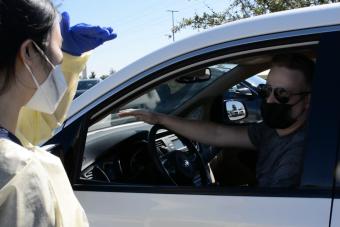 A healthcare worker wearing a face mask, protective gown, and blue gloves speaks to a masked driver in a white car during a drive-through interaction on a sunny day.