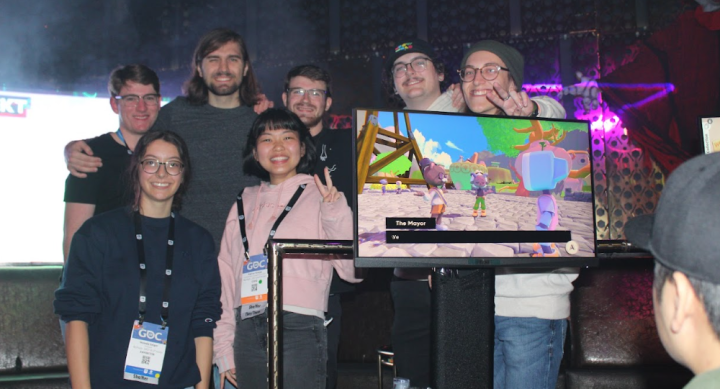 A group of six individuals, pose smiling in front of a display at GDC 2023. They stand behind a monitor showing a colorful game scene with animated characters, including a figure labeled 'The Mayor.' The group includes team members wearing GDC badges and casual attire. The event setting features vibrant lighting and a partially visible red decoration resembling a clown.