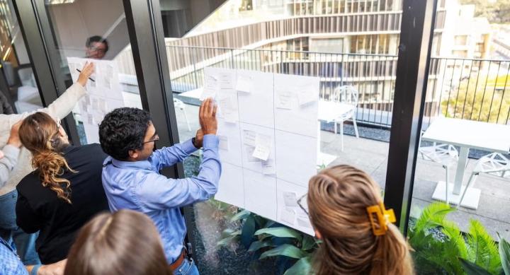 A group of people participating in a design thinking workshop. They are gathered around large sheets of paper with grids and sticky notes, posted on glass windows in a bright, modern building. The background shows an outdoor terrace with white tables and chairs, and a view of contemporary architecture.