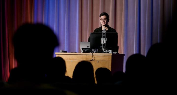 A speaker stands behind a wooden podium, delivering a presentation to an audience in a dimly lit auditorium. The stage backdrop consists of softly lit curtains in shades of blue and red. The audience is visible in the foreground as silhouettes, while the presenter, dressed in black and wearing glasses, speaks into a microphone.