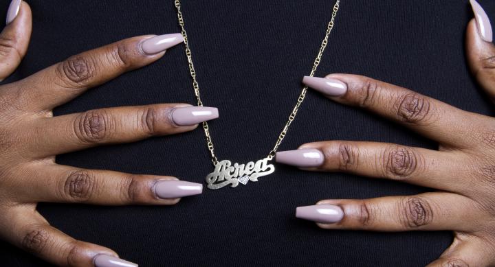 A close-up photograph of a person’s hands with long, glossy, nude-colored nails gently holding a gold nameplate necklace that reads 'Acead' in script font. The necklace has a small heart detail and hangs against a black fabric background.