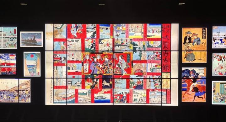 A large digital display wall showcasing a vibrant collage of Japanese woodblock prints. The central composition features a colorful, multi-panel artwork filled with scenes of traditional life, people in elaborate attire, and architectural elements, framed by red borders and Japanese text. Surrounding the center panel are smaller prints, depicting trains, Western visitors, marketplaces, parades, and scenic landscapes. The background is black, emphasizing the illuminated art pieces, which highlight a cultural